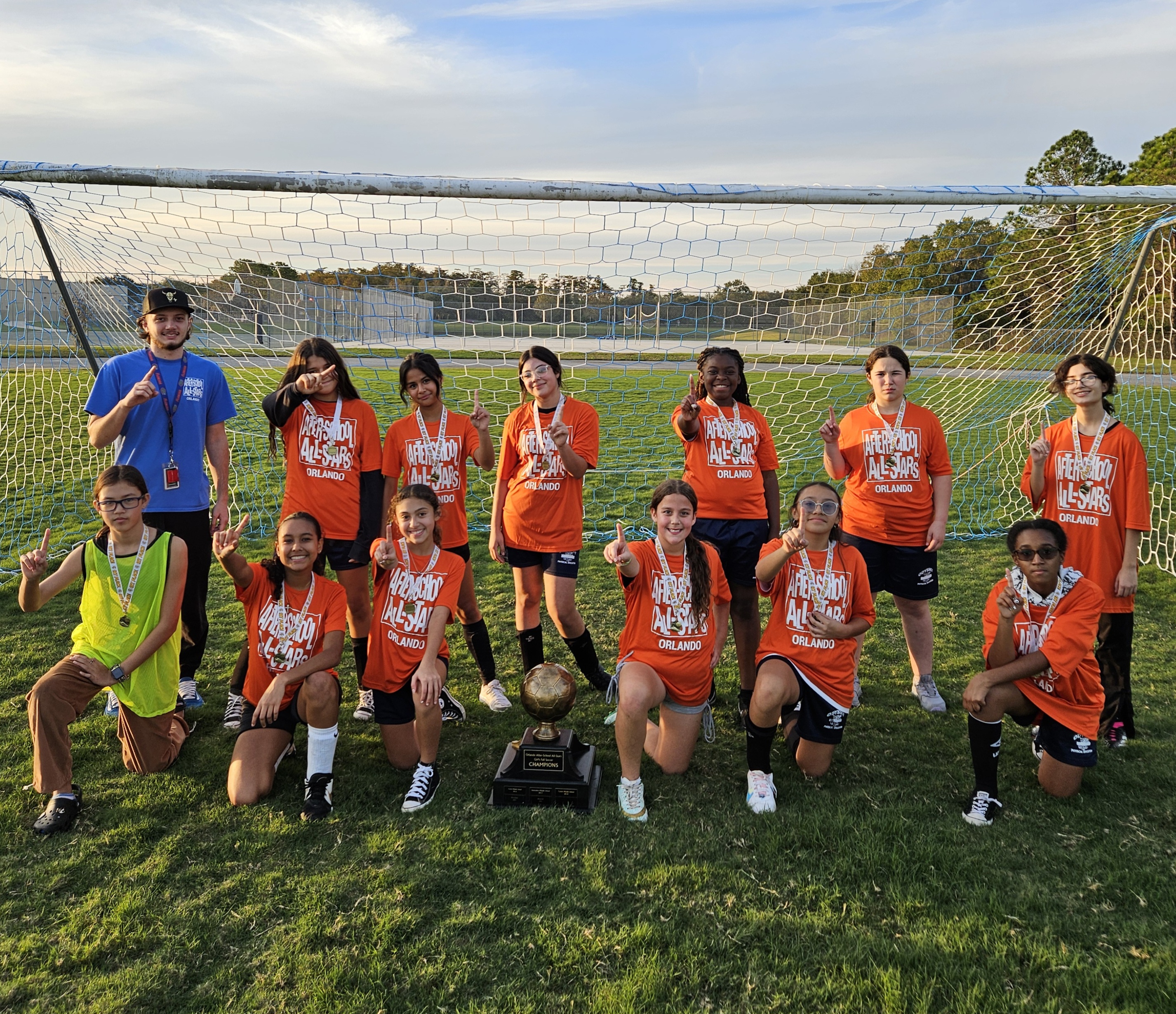 children lined up in front a soccer goal on a field of green grass. Each student is wearing an orange ASAS t-shirt